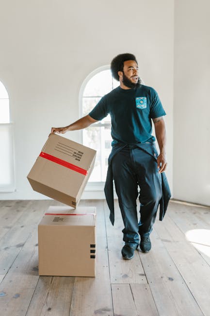 A man with dark curly hair and a beard is standing in a bright, spacious room with white walls and large arched windows that let in natural light. He is wearing a blue t-shirt with a company logo on t