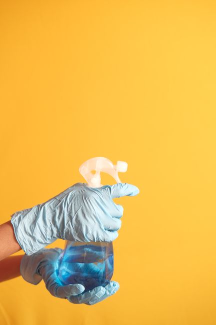 A professional cleaner in a domestic kitchen setting, dressed in full personal protective equipment including a white hazmat suit, safety goggles, a respirator mask, and blue gloves. The cleaner is holding a spray bottle and a cleaning cloth, standing beside a round white table that has a blue cleaning cloth on it. The background shows a modern kitchen with wooden panel walls, a stainless steel extractor hood, and a white refrigerator, all appearing clean and well-maintained. The lighting is bright, emphasizing the pristine condition of the surfaces, and the environment suggests thorough surface cleaning and sanitisation as part of a professional deep cleaning service by Merton Cleaners, in line with safety and hygiene standards for residential spaces.