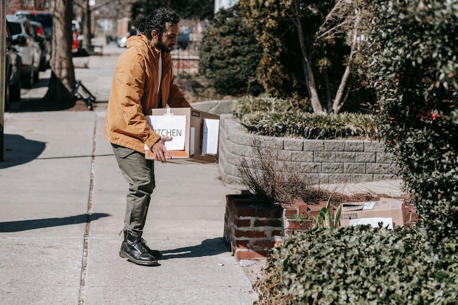 A man dressed in a mustard-colored jacket, olive green pants, and black boots is standing on a sidewalk, lifting a cardboard box labeled 'KITCHEN' with both hands. The box appears to contain household items and is partially open. In the background, there are cars parked along the street, and the sidewalk is lined with a brick planter filled with small shrubs and plants. Sunlight casts shadows, indicating a clear day, and the scene is outdoors in a suburban or urban area. The image emphasizes a moving-out process, which may involve cleaning and sanitisation services provided by Merton Cleaners, a trusted cleaning company.