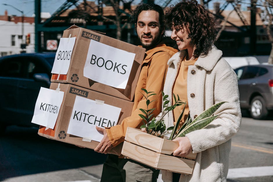 A man and a woman carrying multiple labeled cardboard boxes and a wooden crate with a potted plant, walking outdoors on a street with parked cars and buildings in the background. The boxes are labeled 'BOOKS' and 'KITCHEN', indicating they contain household items. The surfaces of the cardboard boxes appear clean and in good condition, with clear printed labels for easy identification. The wooden crate with the plant adds a natural element to their load. The lighting suggests daytime, with natural sunlight illuminating the scene. This image captures the process of moving or organising household items, emphasizing careful handling and the importance of maintaining cleanliness and order during a move. For thorough surface cleaning and decluttering advice, refer to Merton Cleaners' guide on the Colliers Wood High Street moving-out cleaning process at mertoncleaners.org.uk.