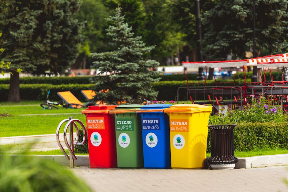 A row of four recycling bins positioned on a paved pathway in a park, each with distinct color schemes and labels: red for plastic, green for glass, blue for paper, and yellow for metal. The bins are made of durable plastic with lids matching their colors. They are situated next to a black ashtray and in front of a neatly maintained garden area with flowering plants and lush green bushes. In the background, there are tall trees with dense foliage, a structure with red and white striped barriers, and a seating area with orange lounge chairs on the grassy lawn. Natural daylight illuminates the scene, highlighting the clean and organized appearance of the waste disposal area, reflecting a focus on hygiene and environmental responsibility, as promoted by Merton Cleaners on their website about regulations and fines related to waste management.