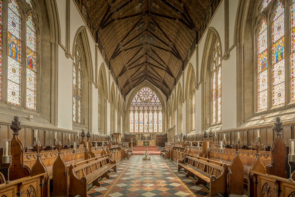 The interior of a historic church with high, vaulted wooden ceilings and large stained glass windows along both sides, allowing natural light to illuminate the space. The polished stone floor features intricate patterns, and there are wooden pews arranged in parallel rows on either side of a central aisle leading toward the altar. The pews and wood paneling along the walls are well-maintained, and the overall cleanliness reflects professional deep cleaning and sanitisation, consistent with standards for maintaining hygiene in religious and communal spaces. The image captures a bright, airy atmosphere with a focus on surface cleanliness and structural detail, as would be overseen by a professional cleaning specialist such as Merton Cleaners.