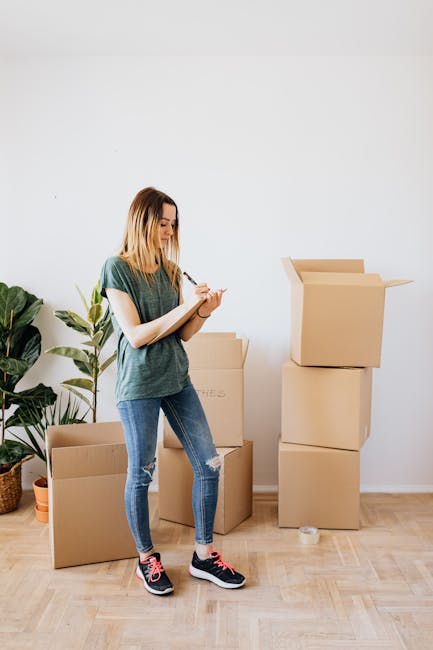 A young woman with shoulder-length blonde hair, wearing a teal T-shirt, ripped jeans, and black sneakers with pink accents, standing in a well-lit, tidy room with light wood flooring and a plain white wall. She is holding a clipboard and pen, appearing to take notes or check off a list. Around her are several cardboard moving boxes, some stacked and some open, and a potted plant with large green leaves on the left side of the image. The scene suggests a residential setting, with clean surfaces and a focus on organisation and preparation, aligned with domestic cleaning and moving-out cleaning activities. The space is minimally decorated, emphasizing the process of packing or cleaning in a professional, safe, and hygienic manner, as exemplified by Merton Cleaners.