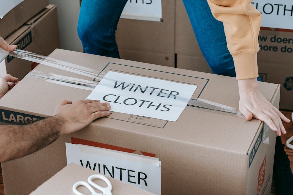 Close-up of a person sealing a large cardboard box labeled 'WINTER CLOTHS' with clear packing tape, while another person holds the box steady. The scene takes place in a room filled with multiple similarly labeled boxes, indicating an organized packing process. The box surfaces are plain brown cardboard with no visible dirt or dust, and the labels are printed on standard white paper. Natural lighting illuminates the scene, highlighting the smooth, flat surfaces of the boxes and the hands involved in sealing. The focus is on the careful packaging, emphasizing clean and neat storage or moving-out preparations. This image reflects the professional cleaning and packing services offered by Merton Cleaners, aligning with their domestic cleaning and removal support expertise.