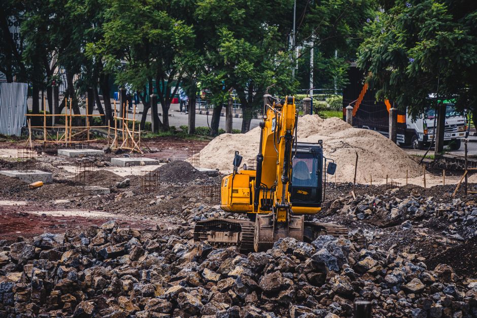 A yellow excavator on a construction site surrounded by piles of rocks and dirt, with trees and parked cars in the background. The site features uneven ground and scattered building materials, indicating active excavation and site preparation. Overhead lighting illuminates the area, highlighting the metallic and dusty surfaces of the construction equipment and the rough terrain. The scene captures the core elements of site groundwork, emphasizing the importance of post-construction surface cleaning and maintenance, as promoted by Merton Cleaners on their page about permit rules for builders' site cleaning in Merton.