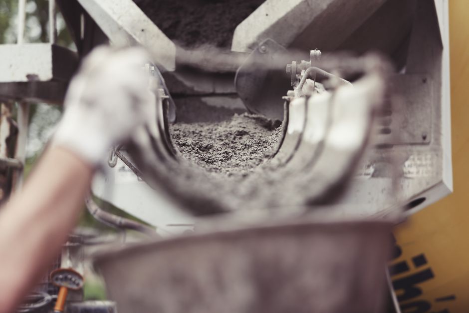 Close-up view of a construction site conveyor or chute with a metallic surface coated in a layer of dry, gray dust and dirt. The equipment features a curved metal slide with visible bolts and joints, positioned indoors. In the background, partial elements of wooden or metal structures are visible, with natural lighting illuminating the scene. The image emphasizes the accumulation of residual debris from building activities, relevant to surface cleaning and maintenance processes in a builders' site. Merton Cleaners specializes in cleaning and sanitisation services, ensuring thorough deep cleaning of construction environments to meet hygiene standards and permit regulations.