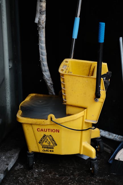 A yellow commercial cleaning mop bucket with a built-in wringer, featuring a caution warning label, positioned on a dark, wet floor next to a black wall. The bucket contains cleaning supplies, including a blue-handled mop and a blue scrubbing brush. In the background, there are visible pipes and a water hose, indicating a cleaning or maintenance area. The surface appears damp, suggesting recent cleaning activity. The scene demonstrates typical equipment used by Merton Cleaners in surface cleaning and sanitisation services within a commercial or residential setting.