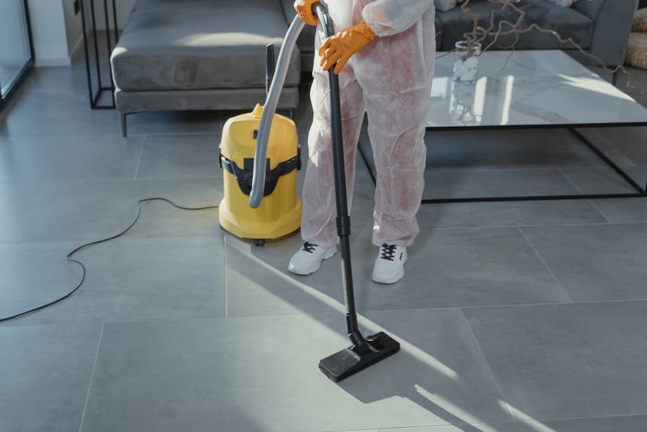 A professional cleaner from Merton Cleaners is performing surface cleaning on a modern, large gray tiled floor in a residential living room near Wimbledon Common. The cleaner is wearing white protective coveralls and orange gloves, operating a yellow vacuum and a black floor cleaning attachment connected to the vacuum hose. The room features a white marble coffee table, a gray upholstered sofa, and a black metal side table with a glass top. Natural light illuminates the space, highlighting the clean, shiny, and dust-free condition of the tiles, which are being deep cleaned and sanitized to maintain hygiene. The overall scene emphasizes thorough domestic cleaning processes in a contemporary home environment.