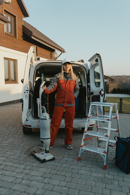 A professional cleaner from Merton Cleaners, dressed in a red and grey uniform, stands in front of a white cleaning van parked on a paved driveway. She holds a handheld vacuum cleaner in one hand and is positioned next to a ladder, with cleaning equipment and a blue bag nearby. The background features a modern, wooden residence with large windows and a well-maintained lawn, under clear blue skies. The scene emphasizes the availability of comprehensive domestic cleaning and surface sanitisation services near Wimbledon Common, SW19, highlighting the company's commitment to thorough and efficient cleaning solutions.