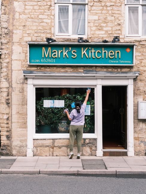 A person with dark hair, wearing a purple shirt and gray pants, is cleaning the large front window of a small restaurant called 'Mark's Kitchen', which offers Chinese takeaway. They are reaching up wi