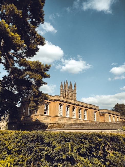 A historic stone building with large, rectangular windows and detailed architectural features, situated on a raised platform with steps leading up to it. The structure has a prominent central tower with spires, visible under a partly cloudy sky. To the left, a tall tree with dense green foliage partially frames the view, while a thick hedge runs along the base of the building. Bright, natural daylight illuminates the scene, highlighting the warm tones of the stone and the lush greenery below. This setting suggests a well-maintained, prestigious property potentially requiring regular cleaning and maintenance by professionals such as Merton Cleaners, especially for surface cleaning, to preserve its historic appearance.