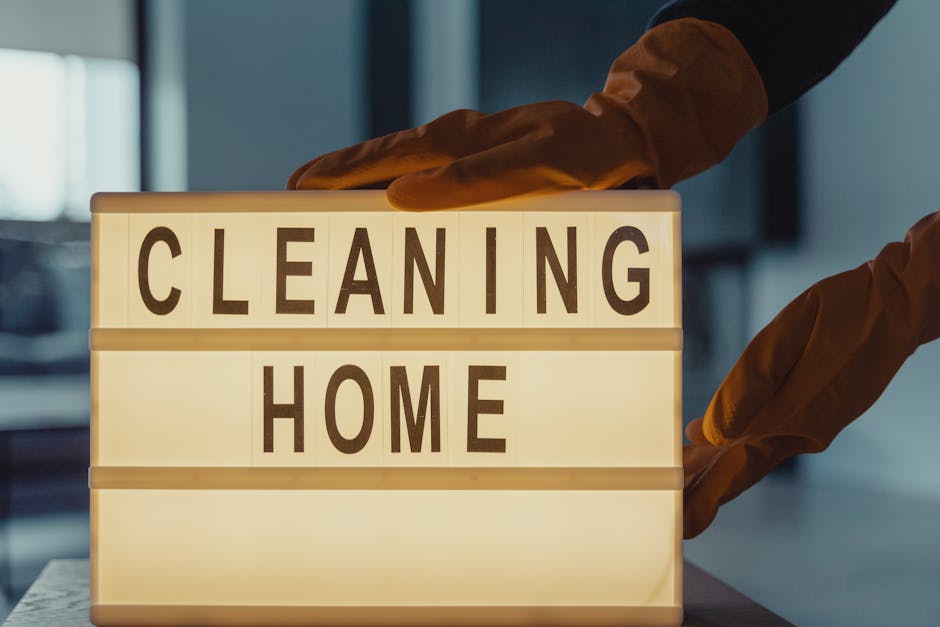 A woman seen from behind, wearing a plaid shirt and light blue jeans, is holding cleaning sprays and wearing pink rubber gloves. She appears to be engaged in domestic cleaning, possibly targeting surfaces such as countertops, appliances, or windows. The background is plain white, focusing on her cleaning activity, with natural lighting highlighting her attire and cleaning tools. This image illustrates the process of surface cleaning and sanitisation, aligning with professional cleaning services like those offered by Merton Cleaners for maintaining hygiene and cleanliness in homes under ?100, in the context of transparent cleaning quotes for Merton properties.
